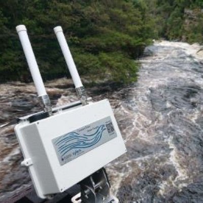 A white camera-looking object with two antennas sits above fast flowing brown water with bushland in the background.